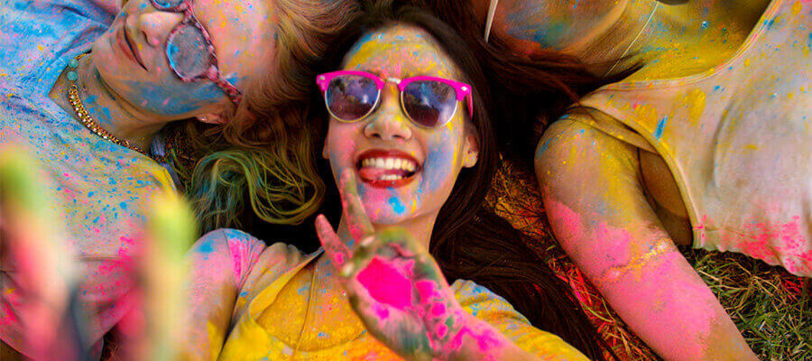 A woman wearing sunglasses, smiling, covered in splashes of multi-coloured paint